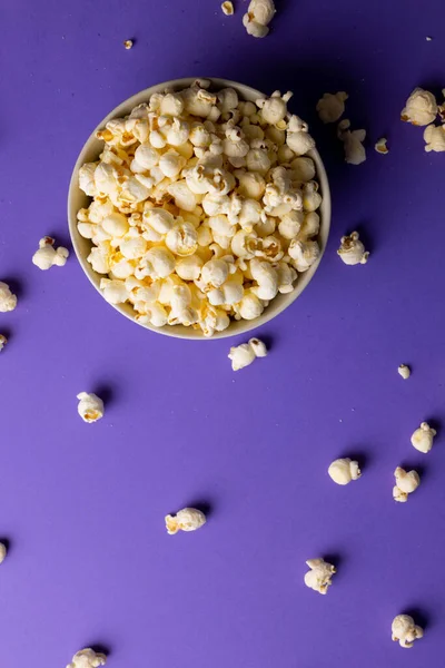 Vertical image of pop corn in bowl lying on violet background. Food, snacks, cinema and american cuisine concept.