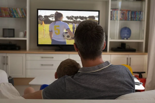 Rear view of father and son sitting at home together watching football match on tv. sports, competition, entertainment and technology concept digital composite image.