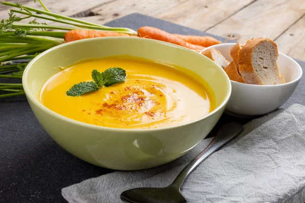 Horizontal image of bowl of carrot soup with garnish, raw carrots and sliced bread on slate. Tasty home cooked food and healthy eating.