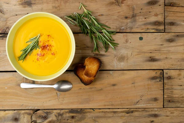 Horizontal image of bowl of carrot soup with rosemary garnish, spoon and toast on wood, copy space. Tasty home cooked food and healthy eating.