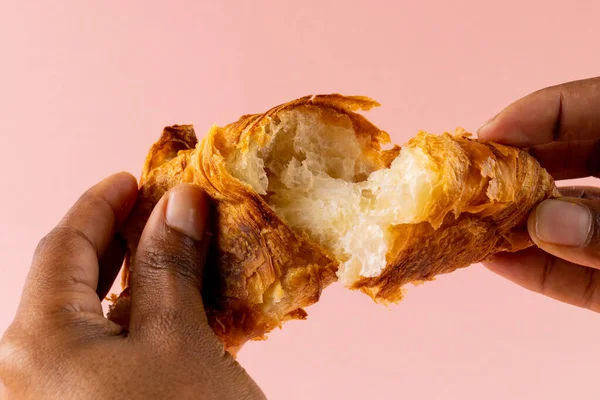 Image of hands of african american man tearing croissants on pink background. Food, confectionery, cakes and traditional french cuisine concept.