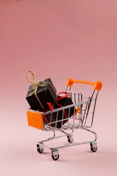 Composition of shopping cart with presents on pink background. Retail, shopping and black friday concept.