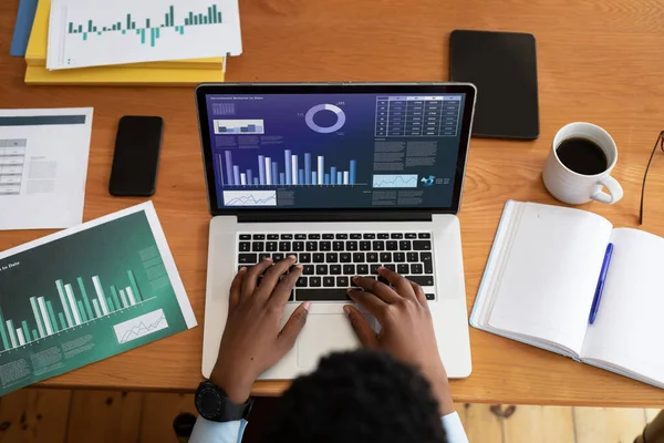 African american businessman sitting at desk using laptop with graph and statistical data on screen. business communication and digital interface technology digital composite image.