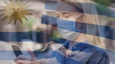 Uruguay bayrağının şehir caddesinde yüz maskesi takan beyaz bir kadının üzerinde dalgalanması. covid 19 salgın konsepti boyunca dijital olarak üretilen küresel sağlık önlemleri.