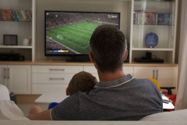 Beyaz tenli baba ve oğul televizyonda futbol maçı izliyorlar. Küresel spor ve kutlama konsepti, dijital kompozit görüntü.