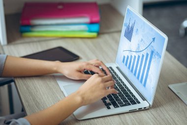 Masadaki beyaz iş kadınının elleri ekranda grafikleri ve verileri olan dizüstü bilgisayarı kullanıyor. İş iletişimi, esnek çalışma, veri ve dijital arayüz kavramı.