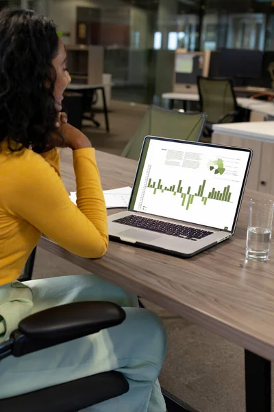 Vertical of biracial businesswoman using laptop in empty office with graph and data on screen. Business communication, flexible working, data, inclusivity and digital interface concept.