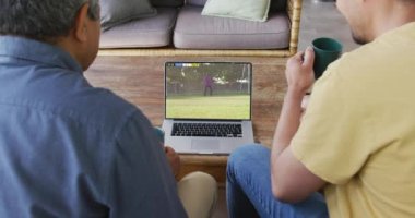 Çift ırklı baba ve oğul ekranda maç yapan çeşitli erkek futbolcularla dizüstü bilgisayar izliyorlar. Küresel spor kavramı, dijital kompozit video.