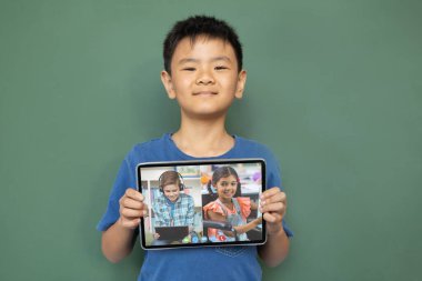 Gülümseyen Asyalı çocuk ekranda çeşitli gülümseyen lise öğrencileriyle video çağrısı için tablet tutuyor. iletişim teknolojisi ve çevrimiçi eğitim, dijital kompozit resim.