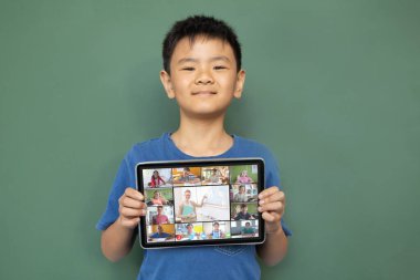 Gülümseyen Asyalı çocuk ekranda çeşitli gülümseyen lise öğrencileriyle video çağrısı için tablet tutuyor. iletişim teknolojisi ve çevrimiçi eğitim, dijital kompozit resim.