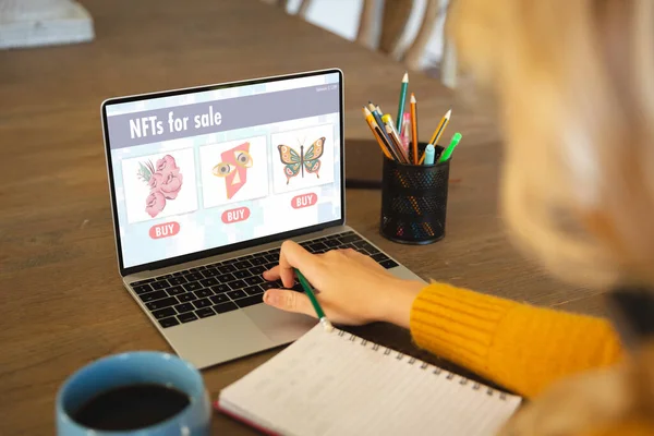 Caucasian businesswoman at table using laptop with nfts for sale on screen. Business communication, flexible working, finance, investment, cryptocurrency and digital interface concept.