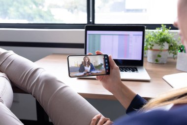 Beyaz kadın akıllı telefon kullanarak masa başında oturuyor ve bayan meslektaşıyla görüntülü görüşme yapıyor. Çevrimiçi toplantı, karantina süresince tecrit altında çalışma.