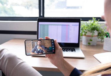 Beyaz bir kadın akıllı telefon kullanarak masa başında oturuyor ve iş arkadaşıyla görüntülü görüşme yapıyor. Çevrimiçi toplantı, karantina süresince tecrit altında çalışma.