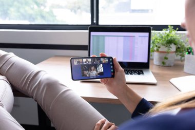 Kafkasyalı iş kadını akıllı telefon kullanarak masada oturuyor ve bir grup meslektaşıyla görüntülü görüşme yapıyor. Modern bir ofiste çalışıyorum..
