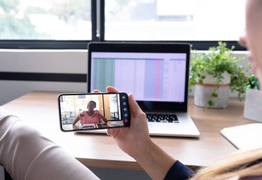 Kafkasyalı iş kadını akıllı telefon kullanarak masa başında oturuyor ve bayan meslektaşıyla görüntülü konuşma yapıyor. Çevrimiçi toplantı, karantina süresince tecrit altında çalışma.