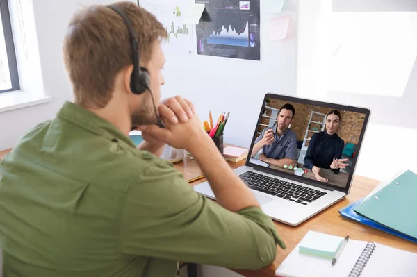 Caucasian man in office having video call with two colleagues displayed on laptop screen. social distancing communication technology workplace during covid 19 pandemic.