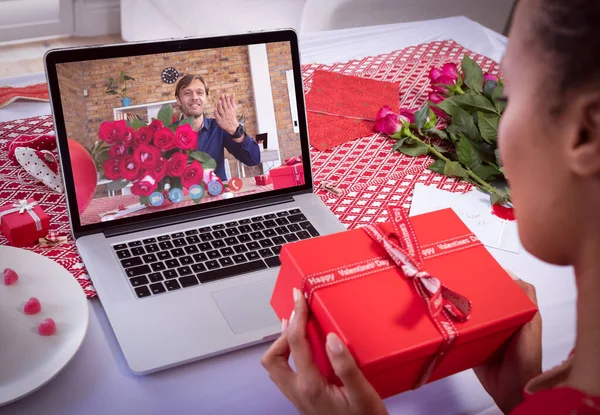 Mixed race woman making valentines date video call holding gift with caucasian man on laptop screen holding flowers. online valentines day communication during quarantine lockdown.