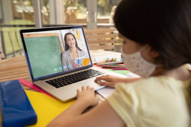 Yüz maskesi takan bir kız öğrenci dizüstü bilgisayarında bayan öğretmenle görüntülü görüşme yapıyor. covid-19 salgını sırasında uzaklardan öğrenme çevrimiçi eğitim