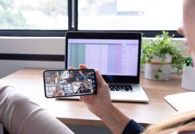 Beyaz kadın akıllı telefon kullanarak masa başında oturuyor ve iş arkadaşlarıyla görüntülü görüşme yapıyor. Çevrimiçi toplantı, karantina süresince tecrit altında çalışma.