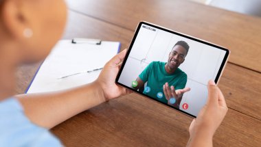 Karışık ırk işkadını masasında oturup tablet kullanıyor ve erkek meslektaşıyla görüntülü konuşma yapıyor. Modern bir ofiste çalışıyorum..