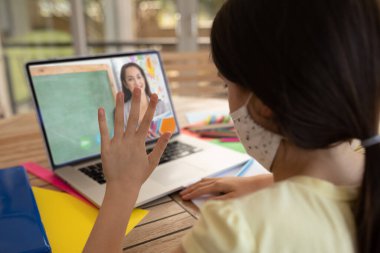 Yüz maskesi takan bir kız öğrenci okulda dizüstü bilgisayarında bayan öğretmenle görüntülü görüşme yapıyor. covid-19 konsepti sırasında uzak öğrenim çevrimiçi eğitim