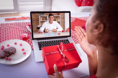 Sevgililer günü videosu çeken melez bir kadın laptoptaki beyaz adama öpücük yolluyor. İkisinde de hediyeler var. Karantina süresince çevrimiçi sevgililer günü iletişimi.