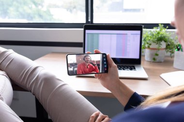 Beyaz iş kadını akıllı telefon kullanarak masasında oturuyor ve erkek meslektaşıyla görüntülü görüşme yapıyor. Modern bir ofiste çalışıyorum..
