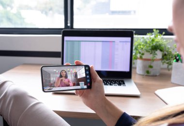 Beyaz bir kadın akıllı telefon kullanarak masa başında oturuyor ve iş arkadaşıyla görüntülü görüşme yapıyor. Çevrimiçi toplantı, karantina süresince tecrit altında çalışma.