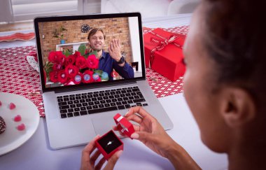 Sevgililer günü kartı hazırlayan melez bir kadın, elinde kırmızı güller tutan beyaz bir adamla görüntülü görüşme yapıyor. Karantina süresince online öneri iletişimi.