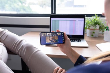 Kafkasyalı iş kadını akıllı telefon kullanarak masada oturuyor ve bir grup meslektaşıyla görüntülü görüşme yapıyor. Modern bir ofiste çalışıyorum..