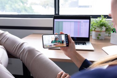 Beyaz kadın öğretmen akıllı telefon kullanarak masada oturuyor ve öğrenciyle online ders alıyor. Karantina süresince evde tecrit altında..
