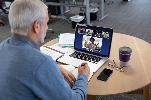 Caucasian man in office having video call with colleagues displayed on laptop screen. social distancing communication technology workplace during covid 19 pandemic.