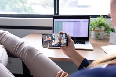 Kafkasyalı iş kadını akıllı telefon kullanarak masada oturuyor ve bir grup meslektaşıyla görüntülü görüşme yapıyor. Modern bir ofiste çalışıyorum..