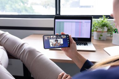 Kafkasyalı iş kadını akıllı telefon kullanarak masada oturuyor ve bir grup meslektaşıyla görüntülü görüşme yapıyor. Modern bir ofiste çalışıyorum..