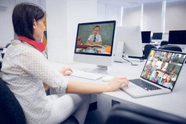 Beyaz kadın öğretmen okul çocuklarıyla video görüşmesinde bilgisayar ve dizüstü bilgisayar kullanıyor. Çevrimiçi eğitim karantina süresince evde tek başına kalıyor..