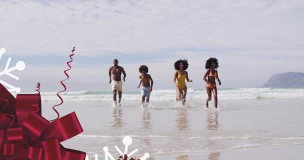 ビーチで走る幸せなアフリカ系アメリカ人の家族の上に赤い弓のアニメーション クリスマス お祝い 家族の時間のコンセプトデジタル生成ビデオ — ストック動画