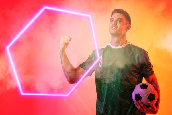 在霓虹灯的背景下 白人男性球员手握足球球 握紧拳头在灯火通明的六边形上 数字合成 霓虹灯 烟雾和抽象概念 — 图库照片