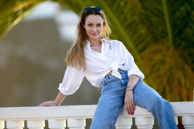 A young beautiful woman in a fashionable dress is sitting on a white fence in the city.