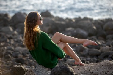 Uzun saçlı model kayalık bir plajda oturuyor. Yeşil kazaklı ve uzun bacaklı güzel bir genç kız deniz dalgalarını izliyor..