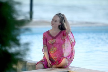 A charming, graceful young blonde woman in a summer dress is sitting on the edge of the swimming pool