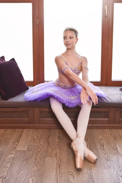 窓のそばに座って プロのバレエダンサーになることを夢見ているパフォーマンス紫色のドレスで美しい小さなバレリーナの完全な長さの肖像画 — ストック写真