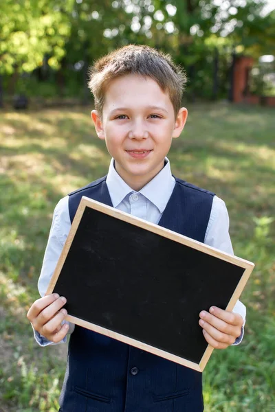 Retrato de un colegial sosteniendo una pequeña pizarra para copiar espacio, sobre un fondo borroso de árboles verdes. — Foto de Stock