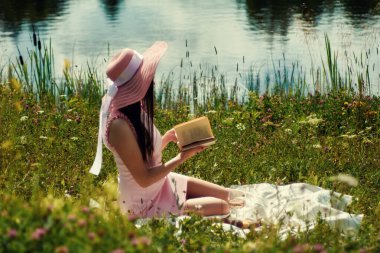 Pembe hasır şapkalı genç bir kadın nehir kenarında oturmuş kitap okuyor..