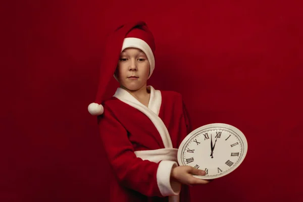 Um menino em um traje de Papai Noel segura um relógio branco em um fundo vermelho. — Fotografia de Stock