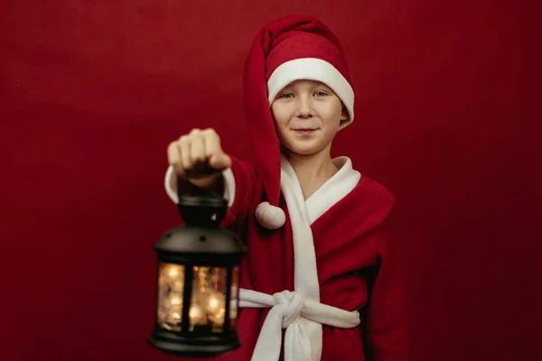 Um menino em um traje de Papai Noel segura uma lanterna de Natal no comprimento dos braços. — Fotografia de Stock