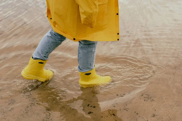 Vidám fiatal nő sárga esőkabátban és sárga csizmában belép a tóba a lábával.. — Stock Fotó
