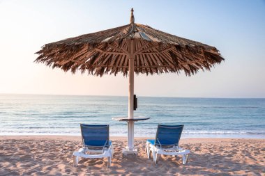 Calm relaxing travel background with neutral colors. Two lounge chairs and palm beach umbrella on sandy sea coast in early morning. Copy space for your text. Luxury holiday vacation on the ocean.