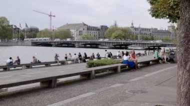 14.08.2021 Zürih Bellevue Gölü gölünde pek çok insan gölde oturur ve bulutlu bir günde hareket halindeki tramvayla Quai Köprüsü 'nün arka planındaki sakin atmosferin tadını çıkarır.
