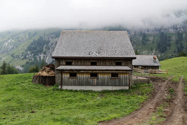 左側に肥料山のある木造住宅6つの窓右側に森の道のある緑の牧草地霧の背景の山の風景 — ストック写真