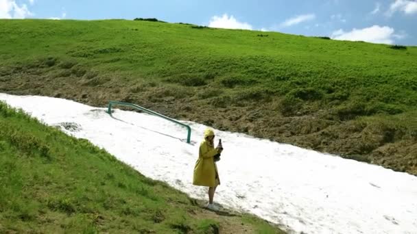 Mulher turista em uma jaqueta amarela brilhante usa um smartphone, tira uma foto contra o fundo de uma montanha coberta de neve em um dia de verão. Turismo e conceito de turismo — Vídeo de Stock